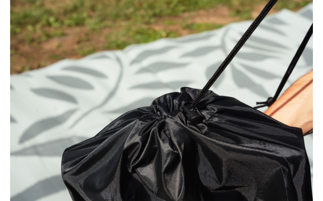 Bolsa negra con cordón sobre una manta clara con estampado de hojas, pie desnudo parcial a la derecha, césped al fondo