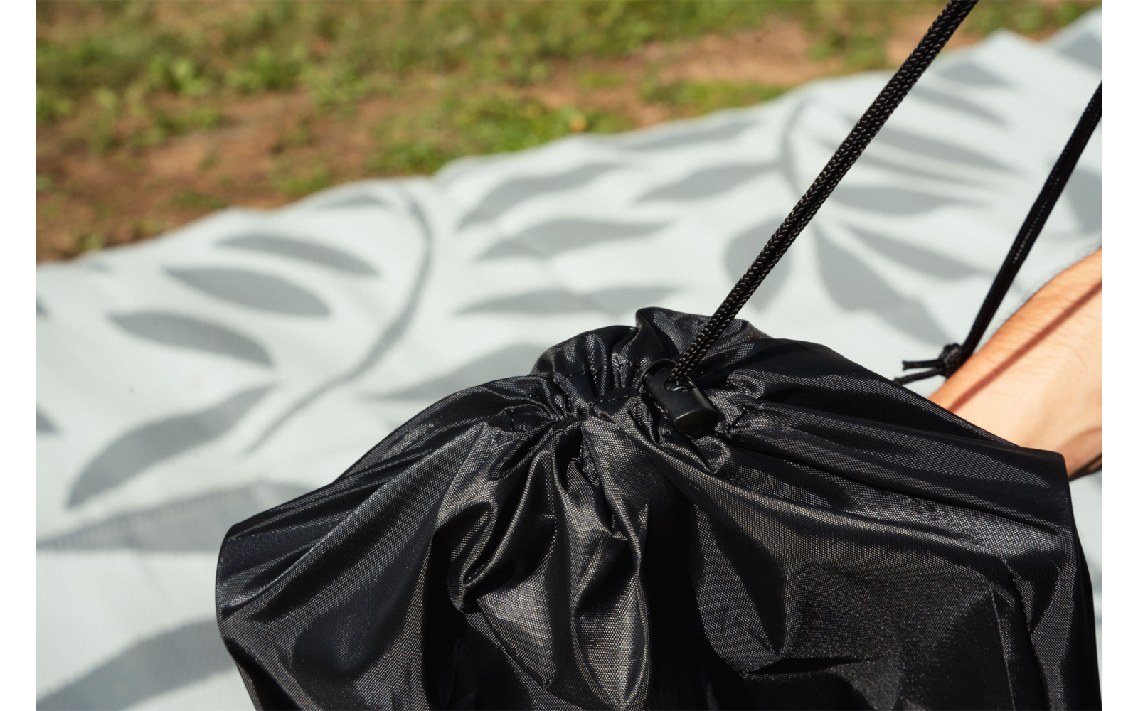 Bolsa negra con cordón sobre una manta clara con estampado de hojas, pie desnudo parcial a la derecha, césped al fondo