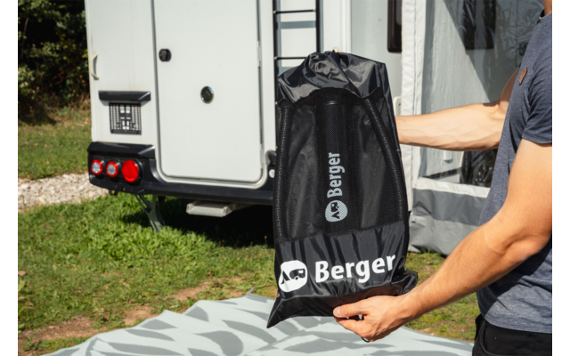 Brazo de una persona sosteniendo una bolsa negra con la inscripción 'Berger' frente a una caravana; césped y alfombra en el suelo
