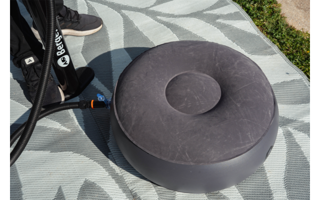 Cojín inflable redondo gris con hendidura central, manguera negra con conector, puntera de zapatilla negra sobre una esterilla gris claro con patrón, hierba en el borde de la imagen