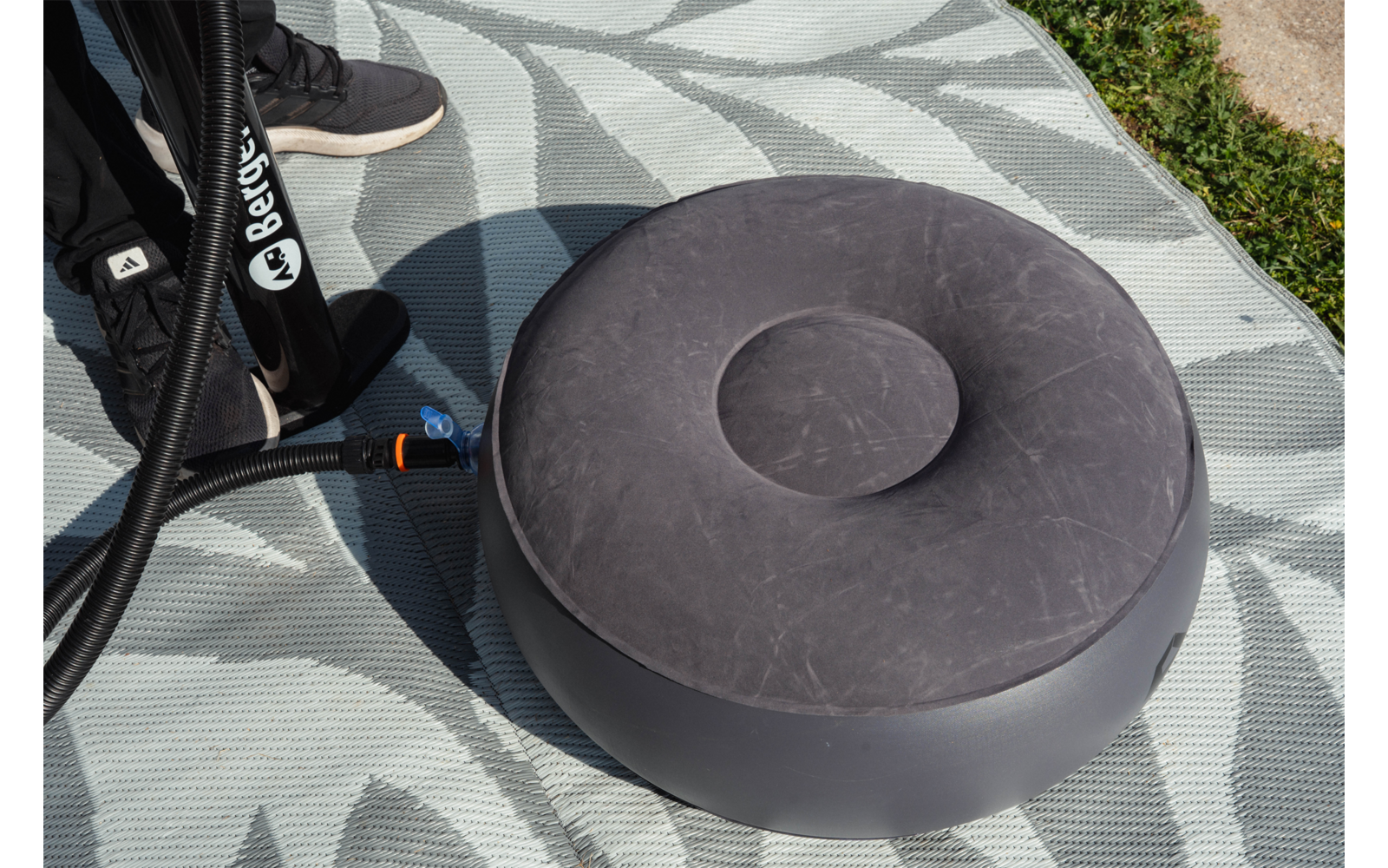 Cojín inflable redondo gris con hendidura central, manguera negra con conector, puntera de zapatilla negra sobre una esterilla gris claro con patrón, hierba en el borde de la imagen