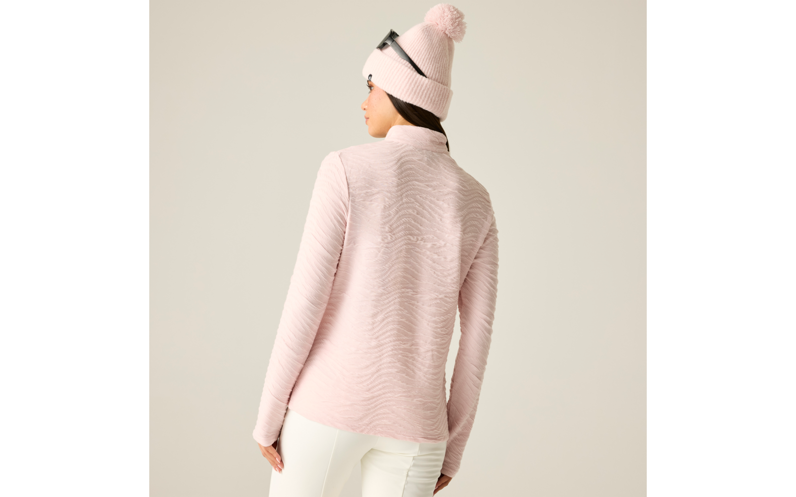 Person von hinten in rosa strukturierter Langarmjacke, weißer Hose, rosa Strickmütze mit Bommel und Brille auf der Mütze, heller Hintergrund