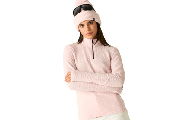 Person mit langen braunen Haaren, rosa Strickmütze mit Bommel und Sonnenbrille auf der Mütze, rosa langärmeliges Oberteil mit Reißverschluss bis zur Brust, verschränkte Arme, weiße Hose, weißer Hintergrund