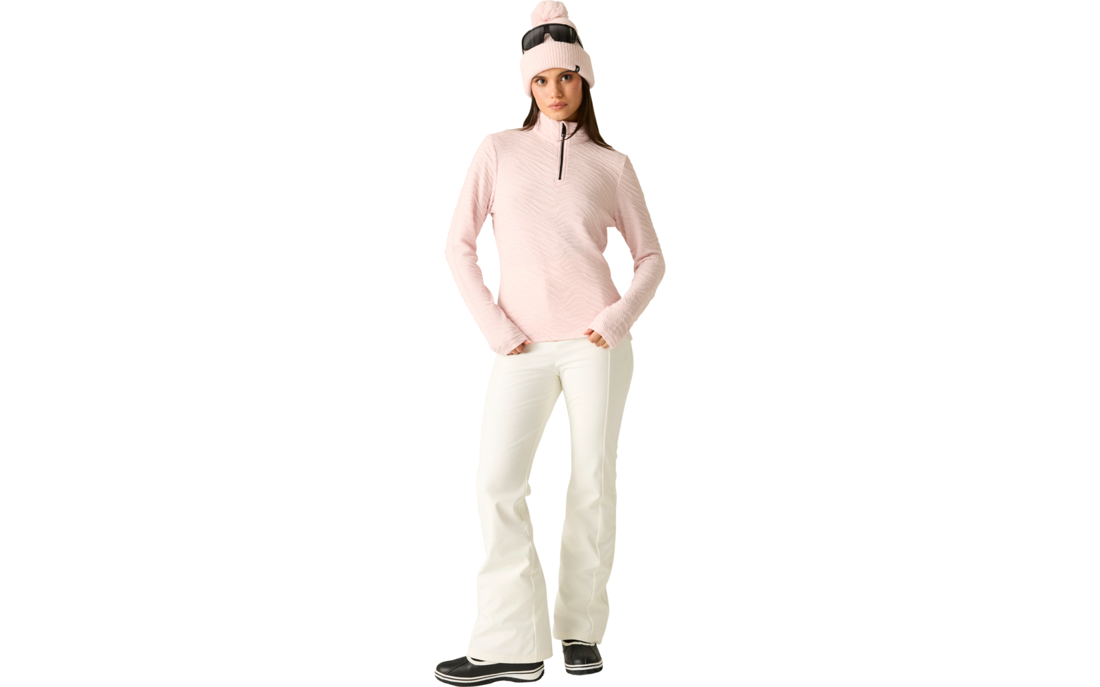 Person mit rosa Strickmütze und Skibrille auf dem Kopf, rosa Fleecepullover mit kurzem Reißverschluss, weiße Skihose und schwarze Stiefel, stehend und frontal