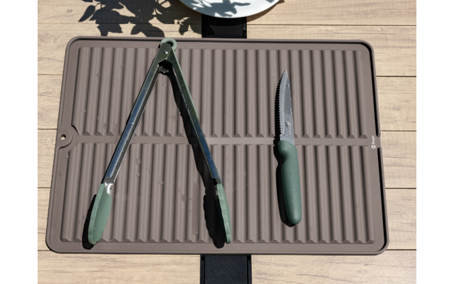 Rechthoekige geribbelde onderlegger op houten oppervlak, een tang met groen handvat links en een gekarteld mes met groen handvat rechts; bovenaan deel van een wit bord en bladvormige schaduwen