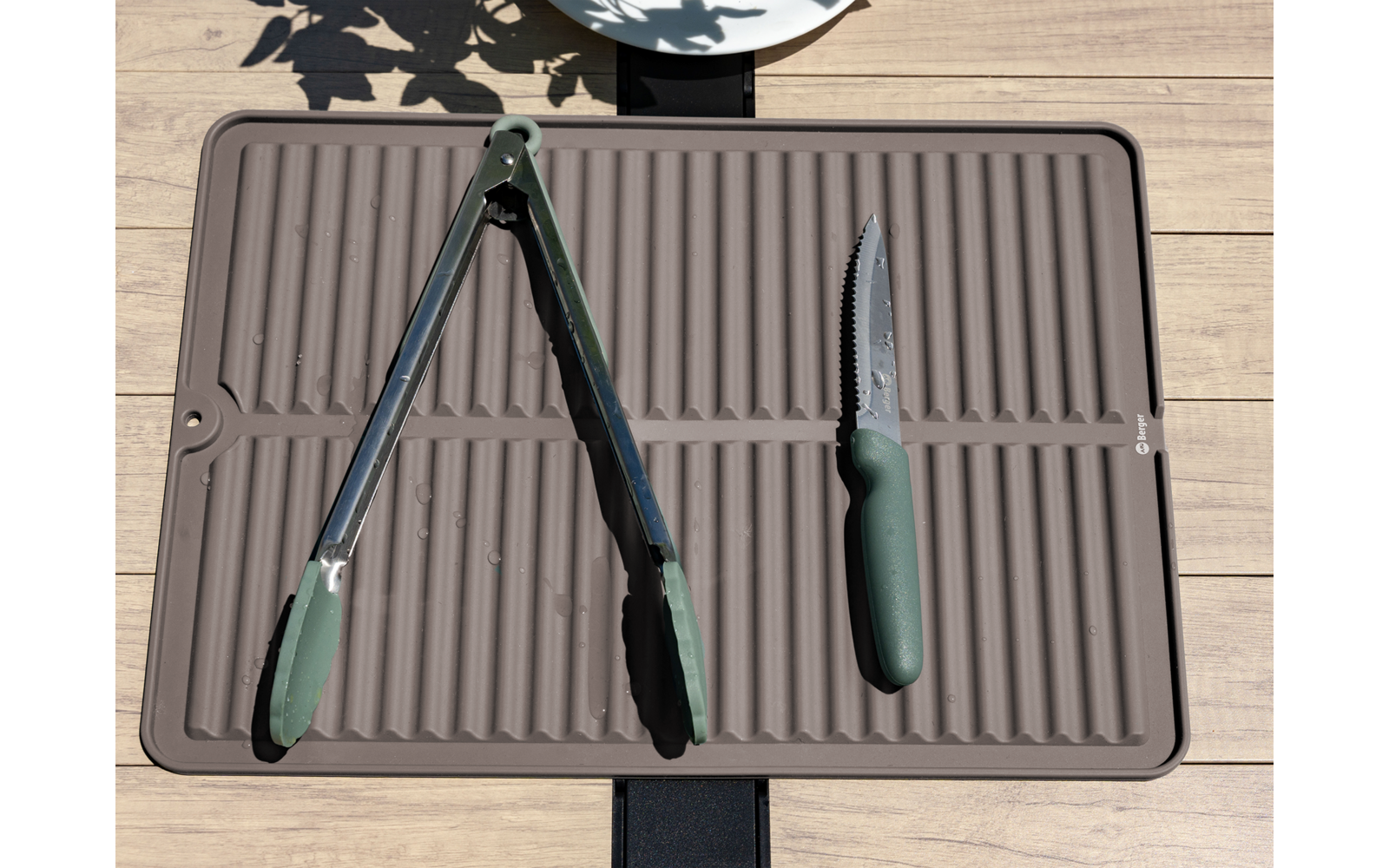 Rechthoekige geribbelde onderlegger op houten oppervlak, een tang met groen handvat links en een gekarteld mes met groen handvat rechts; bovenaan deel van een wit bord en bladvormige schaduwen