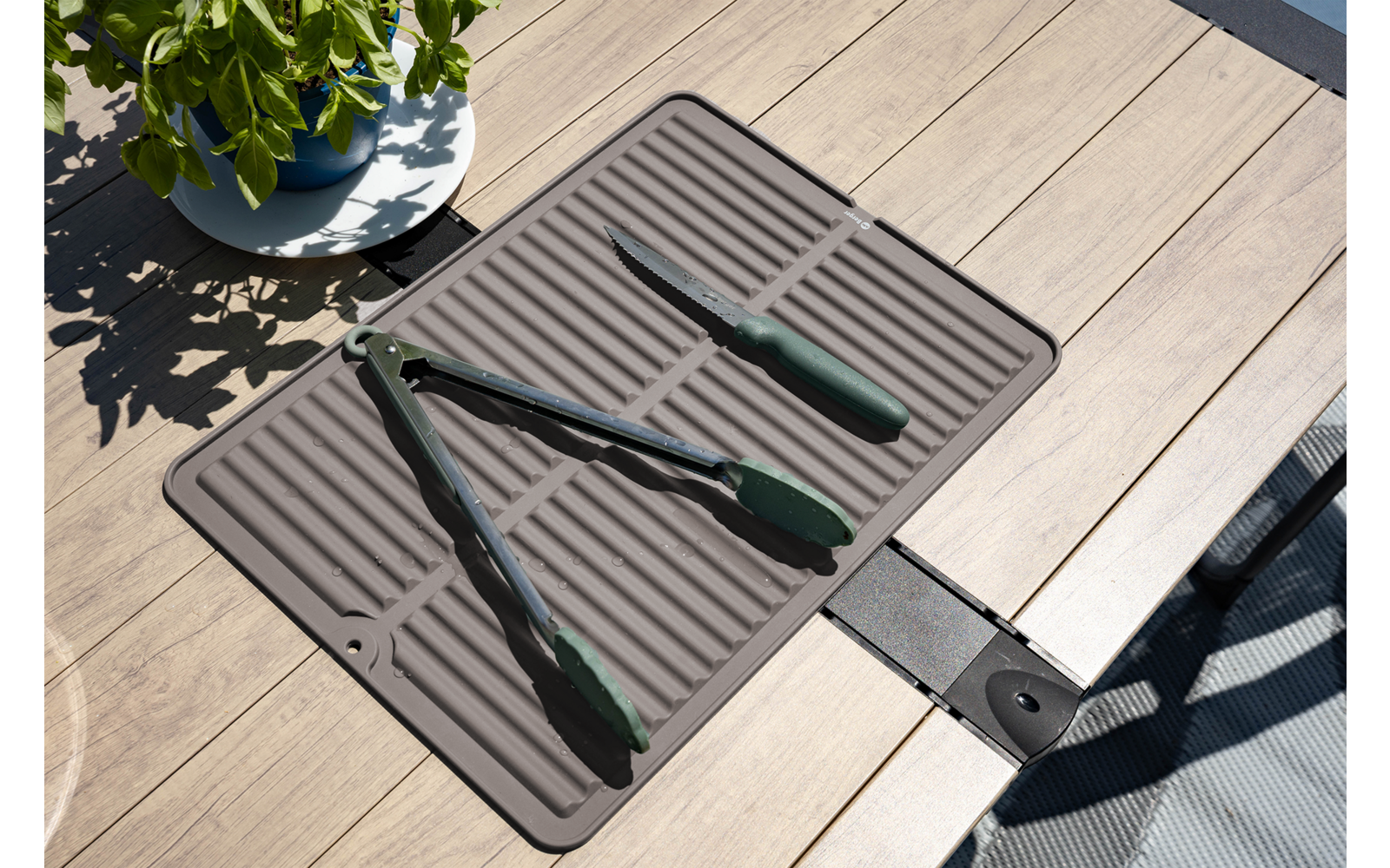 Twee groene tangen en een groene spatel op een grijs geribbeld matje op een houten terras; linksboven een bloempot met bladeren en schaduw