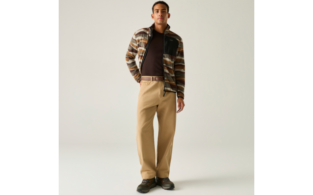 Personne debout devant un fond clair, portant une veste à rayures marron et beige, un haut foncé, un pantalon beige ample avec ceinture et des chaussures foncées