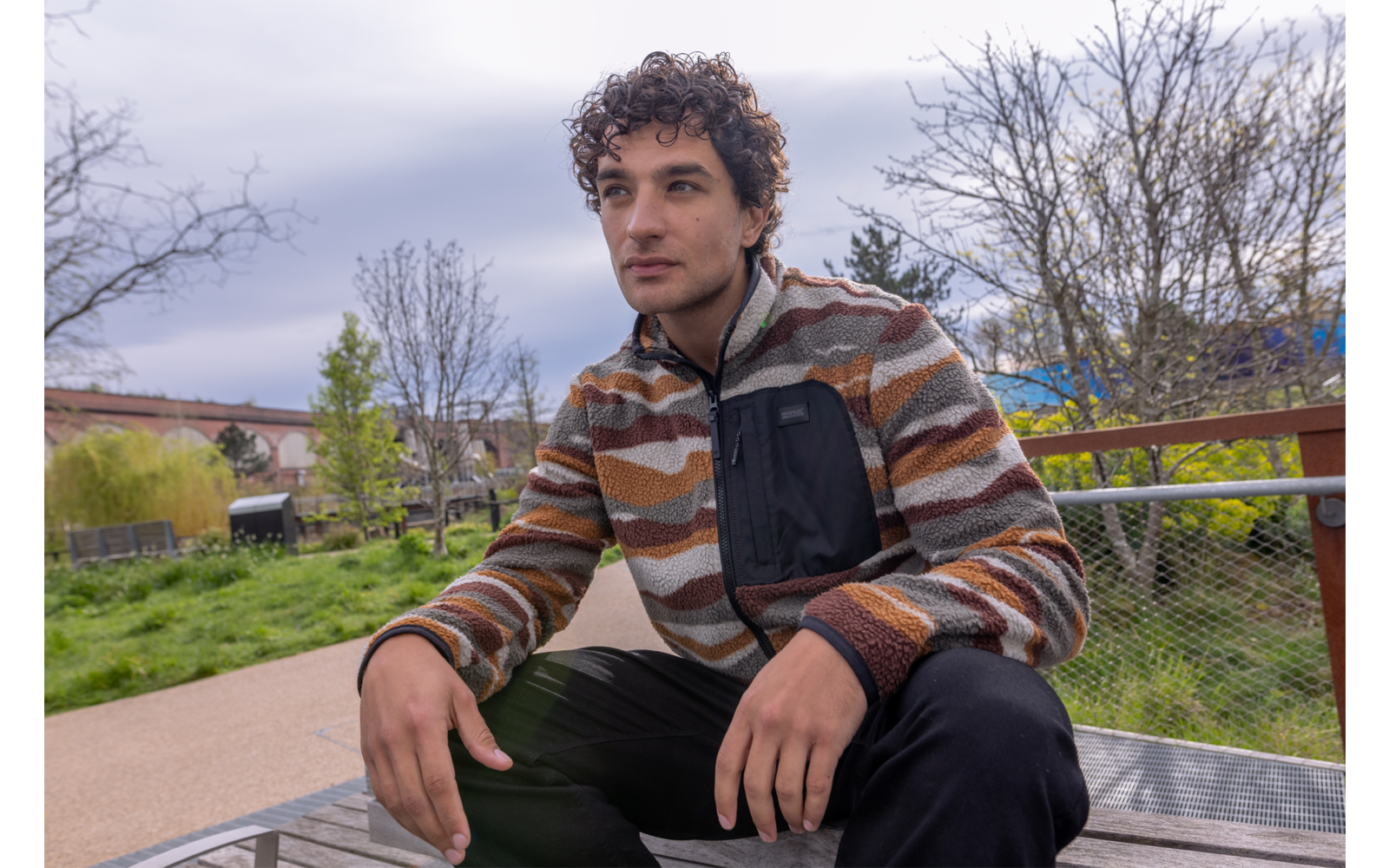 Man met krullend donker haar, draagt een gestreepte fleecejas en zwarte broek, zit op een houten bank in een park, handen op zijn knieën, bomen en bewolkte lucht op de achtergrond