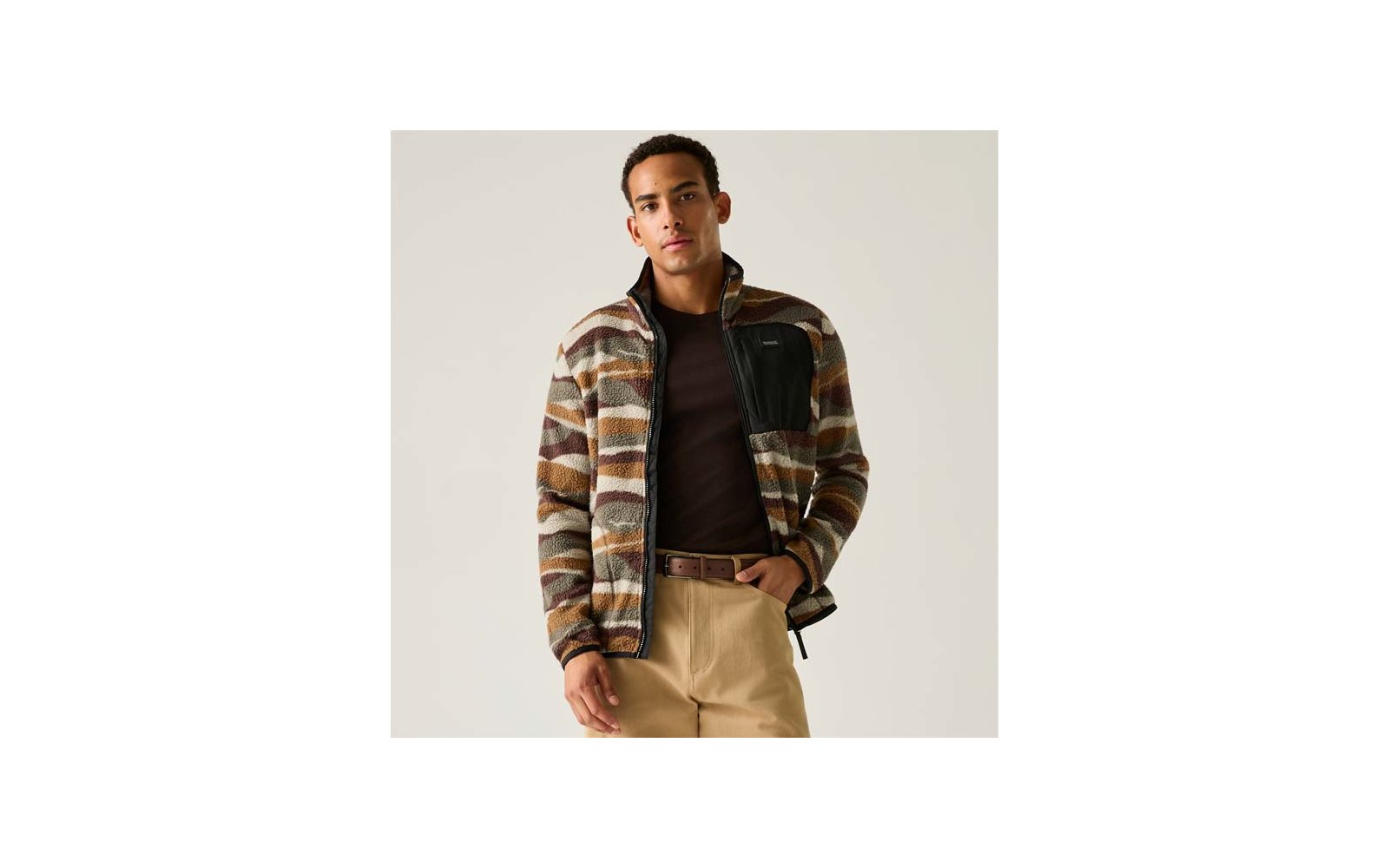 Persoon met een gemêleerd fleecevest met ritssluiting en zwarte borstzak, donker T‑shirt, beige broek en bruine riem, staand voor een lichte achtergrond