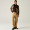 Personne debout devant un fond clair, portant une veste à rayures marron et beige, un haut foncé, un pantalon beige ample avec ceinture et des chaussures foncées