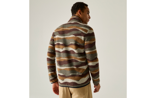 Persoon van achteren met kort krullend haar, draagt een fleecejas met golvende strepen in bruin, olijfgroen en beige en een lichte broek voor een effen lichte achtergrond