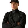 Homme avec casquette noire et pull noir à demi-fermeture éclair, bras croisés, regarde vers la droite