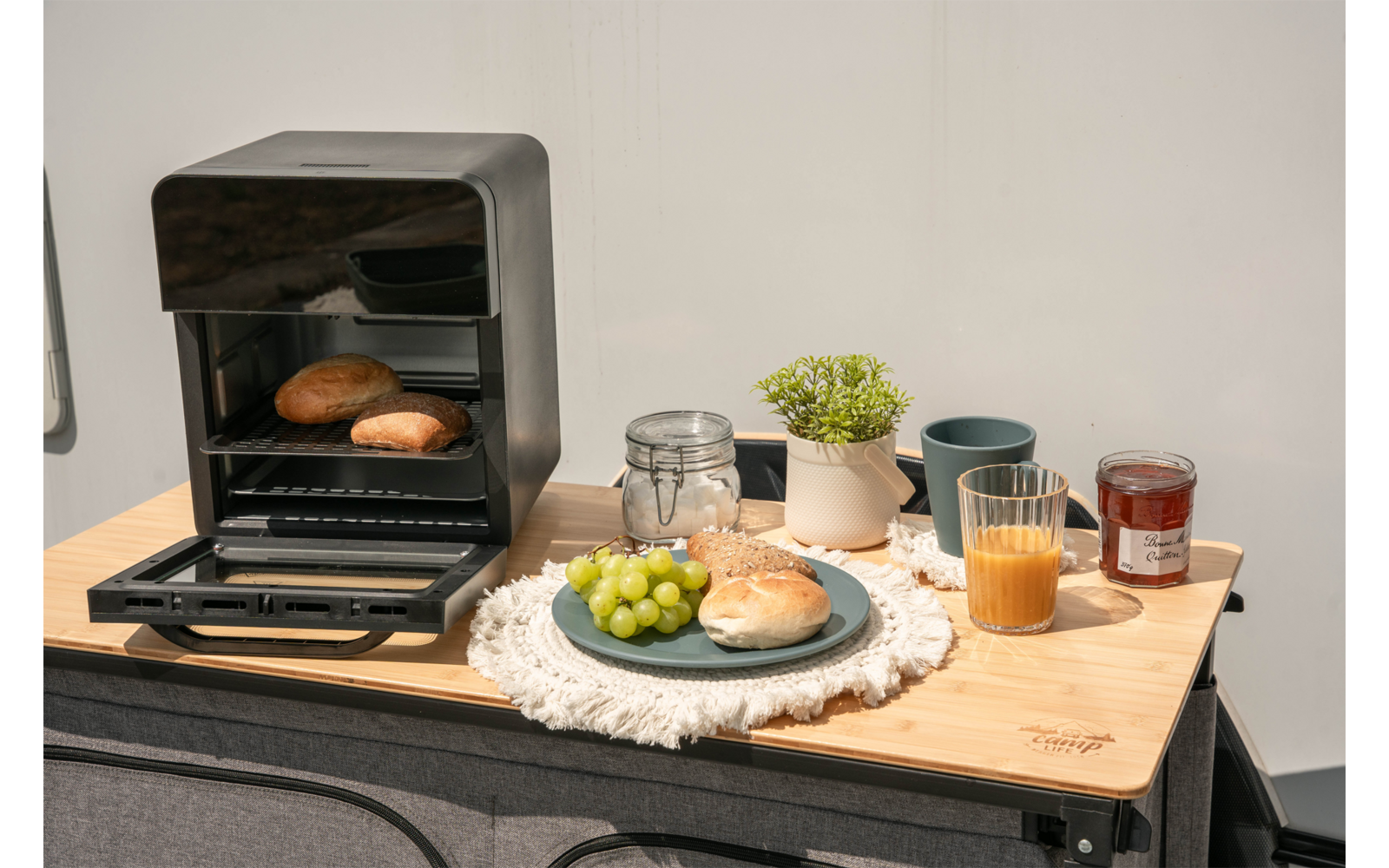 Open mini-oven met twee broodjes binnen; ervoor op een houten oppervlak een bord met een broodje en groene druiven, een glas sinaasappelsap, een pot jam, een weckpot met deksel, een klein bloempotje en een mok