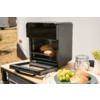 Kleine zwarte tafeloven met open deur en twee broodjes op het rooster; op een houten tafel een bord met groene druiven en een broodje, een glazen pot met beugel, een plantenpot en een doek; caravan en droog gras op de achtergrond
