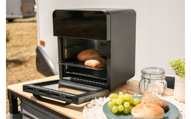 Kleine zwarte tafeloven met open deur en twee broodjes op het rooster; op een houten tafel een bord met groene druiven en een broodje, een glazen pot met beugel, een plantenpot en een doek; caravan en droog gras op de achtergrond