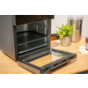 Open toasteroven met geopende glazen deur op een houten werkblad; daarnaast een groene potplant, een roestvrijstalen bus en een pot met deksel