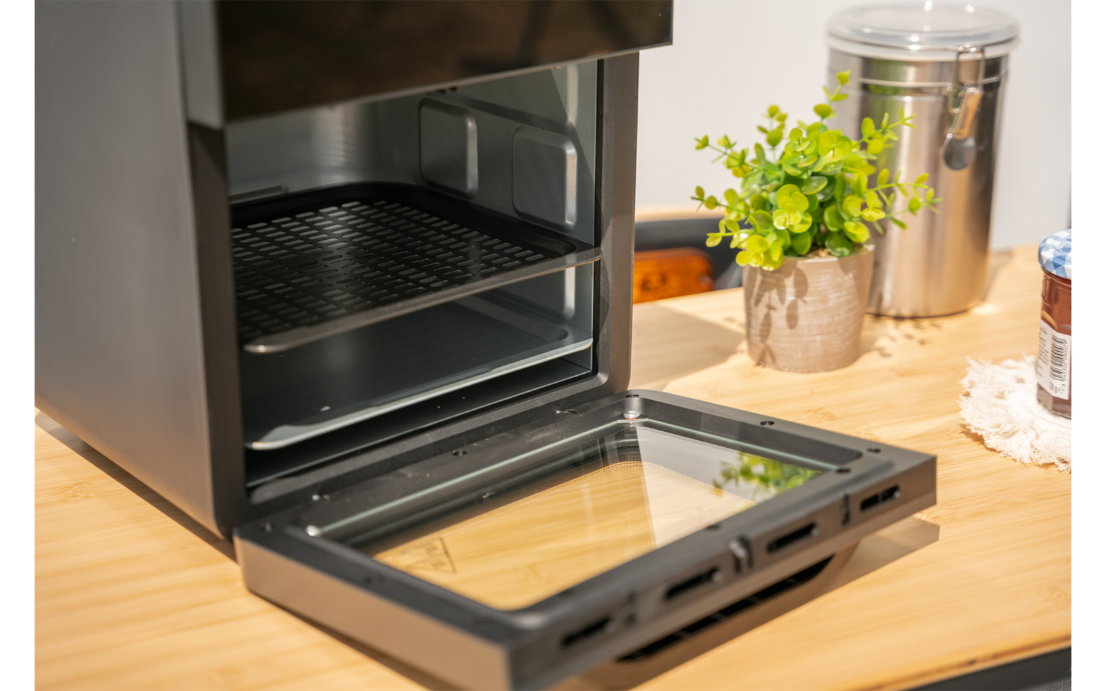 Open toasteroven met geopende glazen deur op een houten werkblad; daarnaast een groene potplant, een roestvrijstalen bus en een pot met deksel