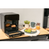 Open zwart mini-oven met twee broodjes erin op een houten blad; daarnaast een bord met groene druiven en een broodje, een glas sinaasappelsap, een grijze mok, een glazen pot met beugel, een klein plantenpotje en een wit doekje