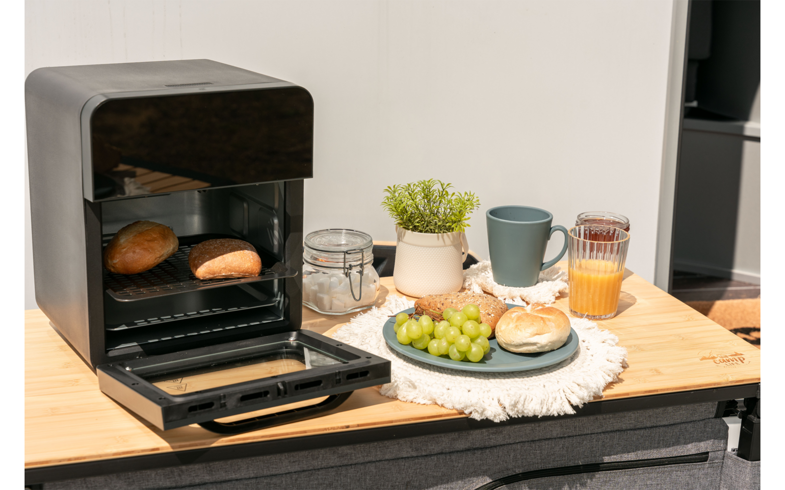 Open zwart mini-oven met twee broodjes erin op een houten blad; daarnaast een bord met groene druiven en een broodje, een glas sinaasappelsap, een grijze mok, een glazen pot met beugel, een klein plantenpotje en een wit doekje