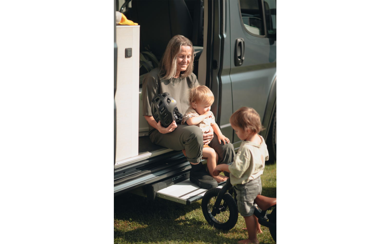 Mujer sentada en la parte trasera abierta de una furgoneta sosteniendo un casco de bicicleta; dos niños pequeños, uno en su regazo, el otro de pie con una bicicleta sin pedales sobre el césped