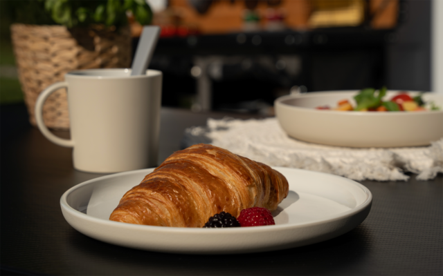 Croissant auf weißem Teller, davor eine Himbeere und eine Brombeere; im Hintergrund eine beige Tasse mit Löffel und eine Schale mit Salat auf dunklem Tisch
