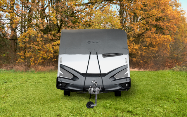 Frontansicht eines Wohnwagens mit grauer Abdeckung oben, Anhängerkupplung in der Mitte, auf grünem Rasen vor herbstlichen Bäumen
