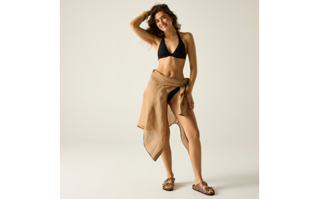 Person steht vor hellem einfarbigem Hintergrund, trägt schwarzes Bikinioberteil, beigen Wickelrock an der Taille gebunden, Sandalen mit Leopardenmuster, eine Hand im Haar, lächelnd