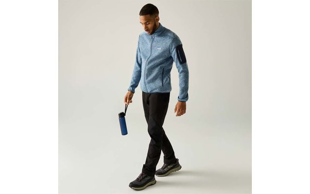 Persoon loopt, draagt een lichtblauwe ritsjas met mouwzak, zwarte broek en zwarte sportschoenen, houdt een blauwe drinkfles in de rechterhand en kijkt omlaag