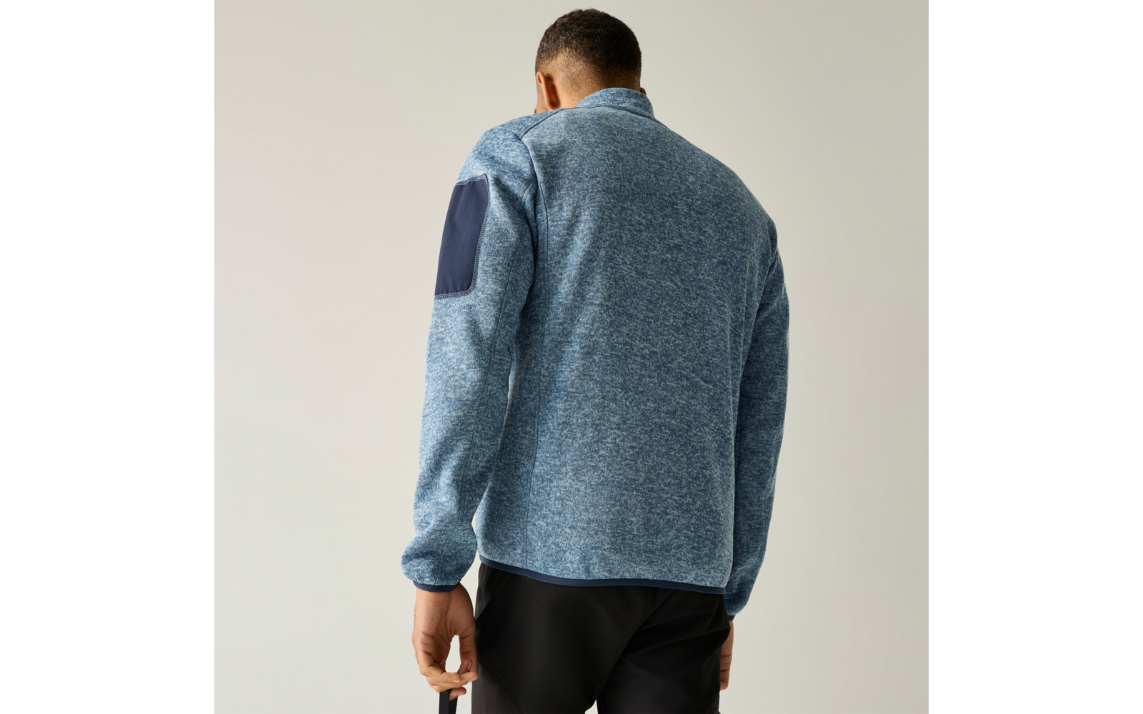Persoon van achteren in lichtblauw gemêleerd fleecejack met donkerblauwe elleboogpatch op de rechtermouw, zwarte broek, neutrale achtergrond