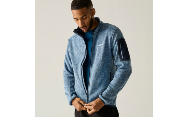 Persoon met kort haar die een lichtblauwe fleecejas met ritssluiting en ritszak op de mouw over een blauw shirt sluit, voor een lichte achtergrond
