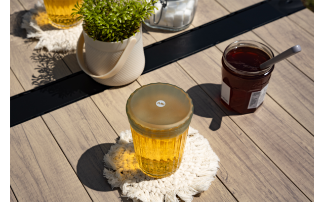 Geribbeld glas met geel drankje op een wit gehaakt onderzetter op een houten tafel; rechts een open pot met rode inhoud en een lepel; daarachter een wit potje met een groene plant