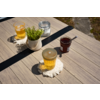 Houten tafel met twee glazen met amberkleurige vloeistof op gehaakte onderzetters, kleine witte pot met groene plant, een gesloten glazen pot en een open jam-pot met lepel; steenachtige ondergrond zichtbaar bij tafelhoek