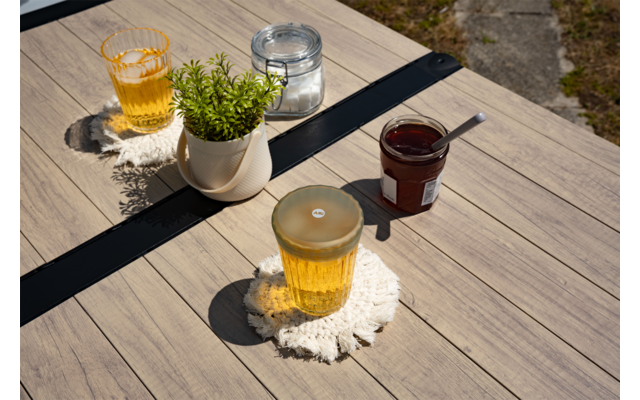Houten tafel met twee glazen met amberkleurige vloeistof op gehaakte onderzetters, kleine witte pot met groene plant, een gesloten glazen pot en een open jam-pot met lepel; steenachtige ondergrond zichtbaar bij tafelhoek