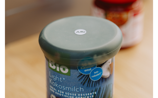Plastic bakje met groene deksel en blauw etiket 'Bio Light Kokosmilch' op een houten oppervlak, onscherpe pot met rood deksel op de achtergrond