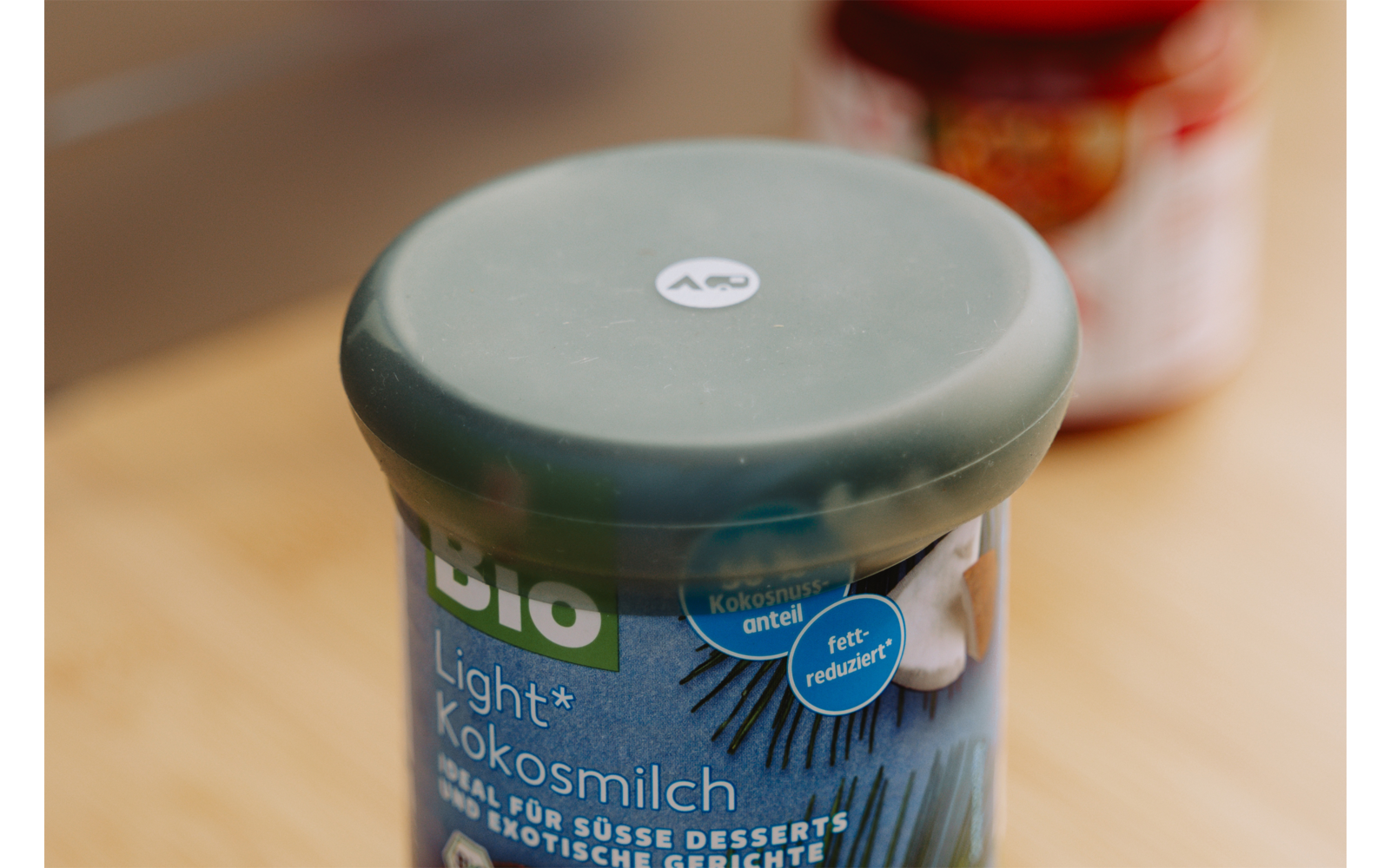 Plastic bakje met groene deksel en blauw etiket 'Bio Light Kokosmilch' op een houten oppervlak, onscherpe pot met rood deksel op de achtergrond