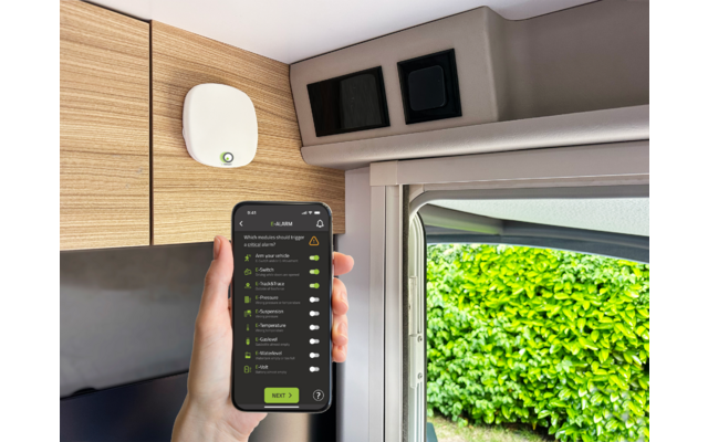 Hand houdt smartphone met een lijst op het scherm; interieur met houten kasten, een wit apparaat aan de wand en geopend raam; groene haag buiten