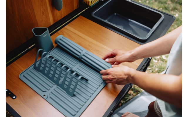 Hände platzieren eine graue Silikon-Abtropfmatte mit Abtropfgestell auf einer Holzarbeitsplatte neben einem schwarzen Waschbecken; links ein grauer Besteckbecher und draußen Gras