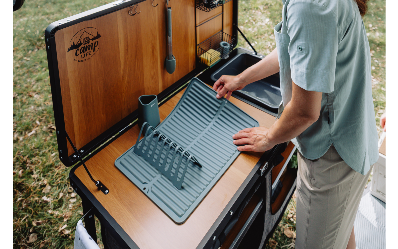 Person stellt Abtropfgestell auf Abtropfmatte neben Spüle einer mobilen Campingküche im Freien