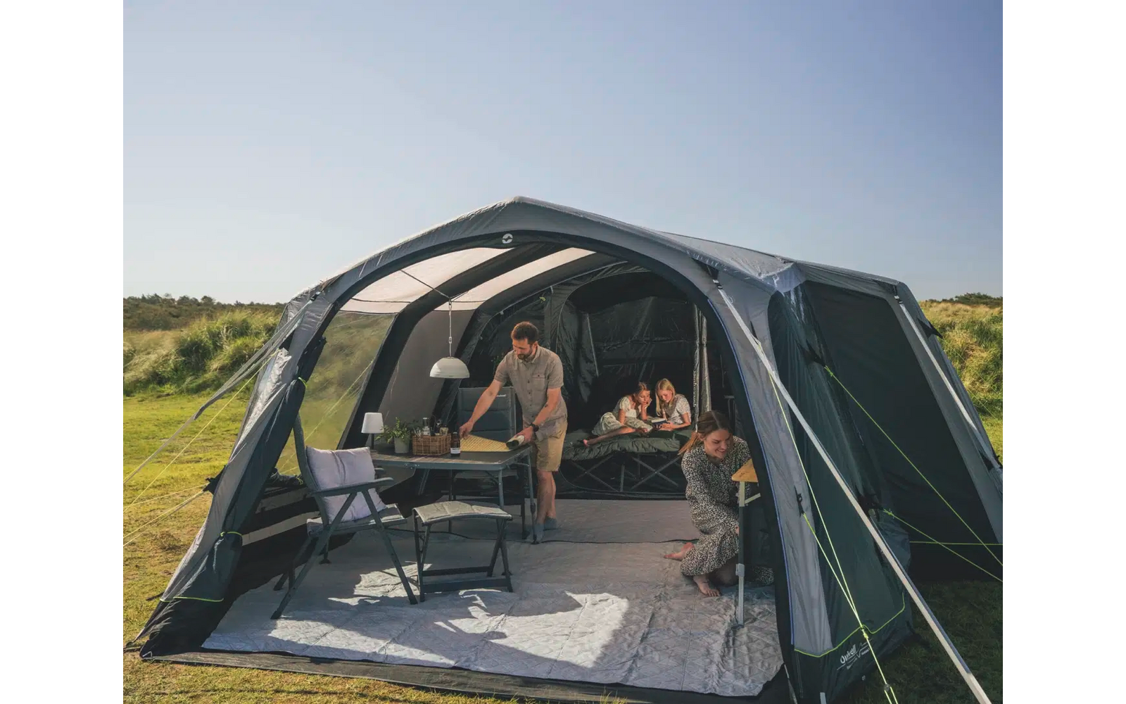 Zwei Personen sitzen in einem großen, offenen Zelt in einer grünen Landschaft mit Wald im Hintergrund