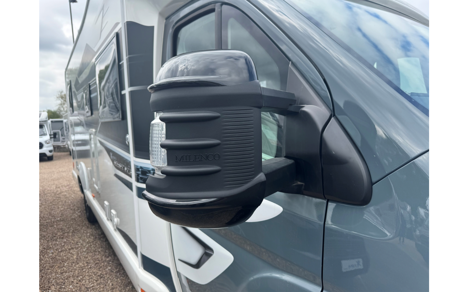 Nahaufnahme eines seitlichen Außenspiegels an einem grauen Wohnmobil mit schwarzer Spiegelabdeckung; weitere Wohnmobile und bewölkter Himmel im Hintergrund