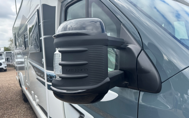 Nahaufnahme eines seitlichen Außenspiegels an einem grauen Wohnmobil mit schwarzer Spiegelabdeckung; weitere Wohnmobile und bewölkter Himmel im Hintergrund