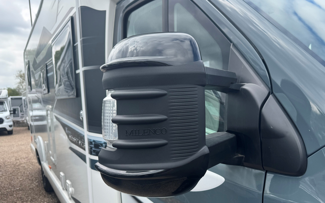 Close-up van een zijspiegel van een grijze camper met een zwarte spiegelkap; andere campers en een bewolkte lucht op de achtergrond