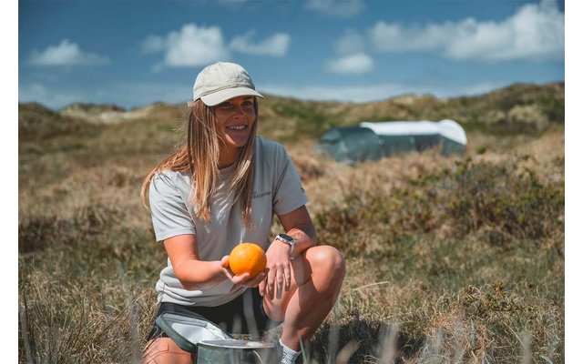Persona sentada sobre una nevera portátil en la hierba sosteniendo una naranja, lleva gorra, camiseta y pantalones cortos; dunas y cielo azul con nubes al fondo
