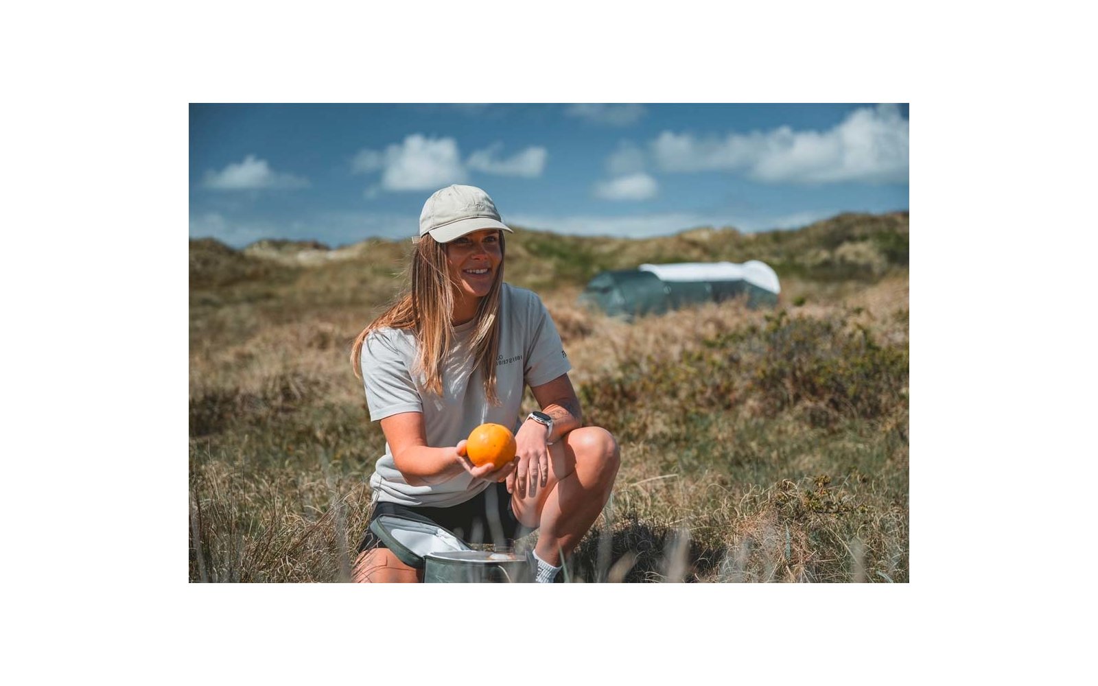 Persona sentada sobre una nevera portátil en la hierba sosteniendo una naranja, lleva gorra, camiseta y pantalones cortos; dunas y cielo azul con nubes al fondo