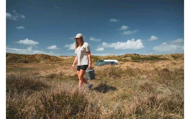 Persona con gorra blanca, camiseta blanca y pantalones cortos negros llevando un cubo metálico, caminando por dunas arenosas con hierba alta; cielo azul con nubes dispersas