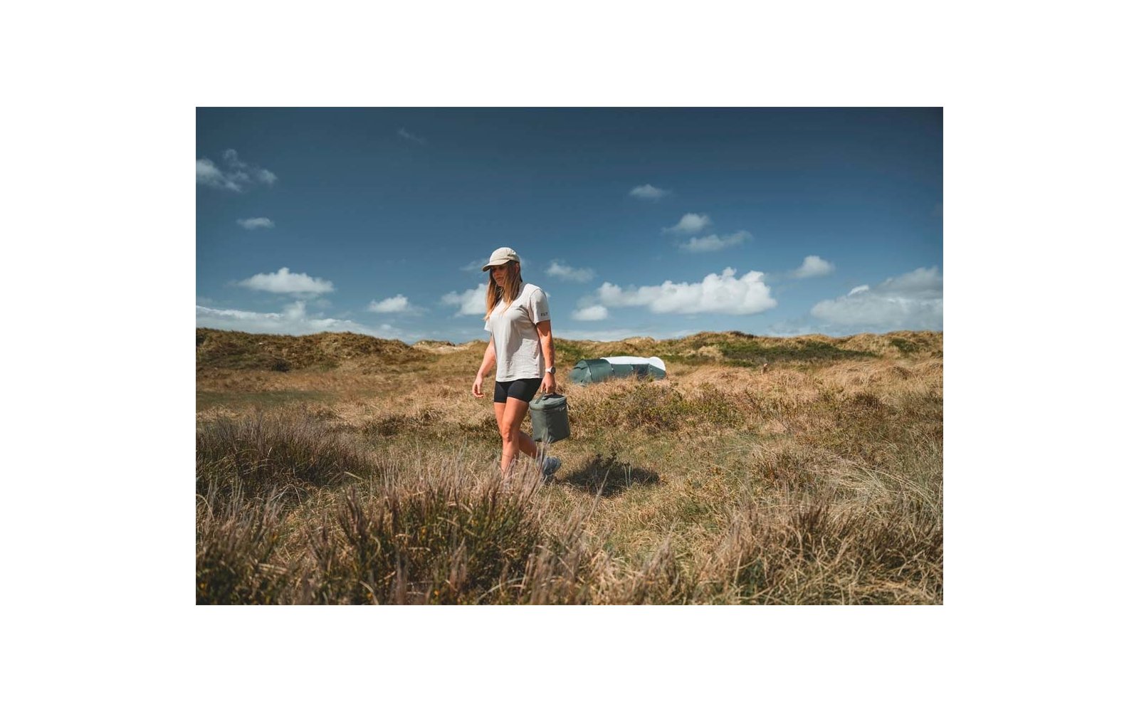 Persona con gorra blanca, camiseta blanca y pantalones cortos negros llevando un cubo metálico, caminando por dunas arenosas con hierba alta; cielo azul con nubes dispersas