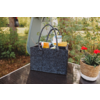 Dunkelgraue Filztasche mit zwei Tragegriffen auf einem glänzenden Tisch, darin mehrere Plastikflaschen mit gelben und orangen Verschlüssen und eine kleine weiß-blaue Schachtel; rechts ein geflochtener Blumentopf mit roten Blüten, im Hintergrund Pflan