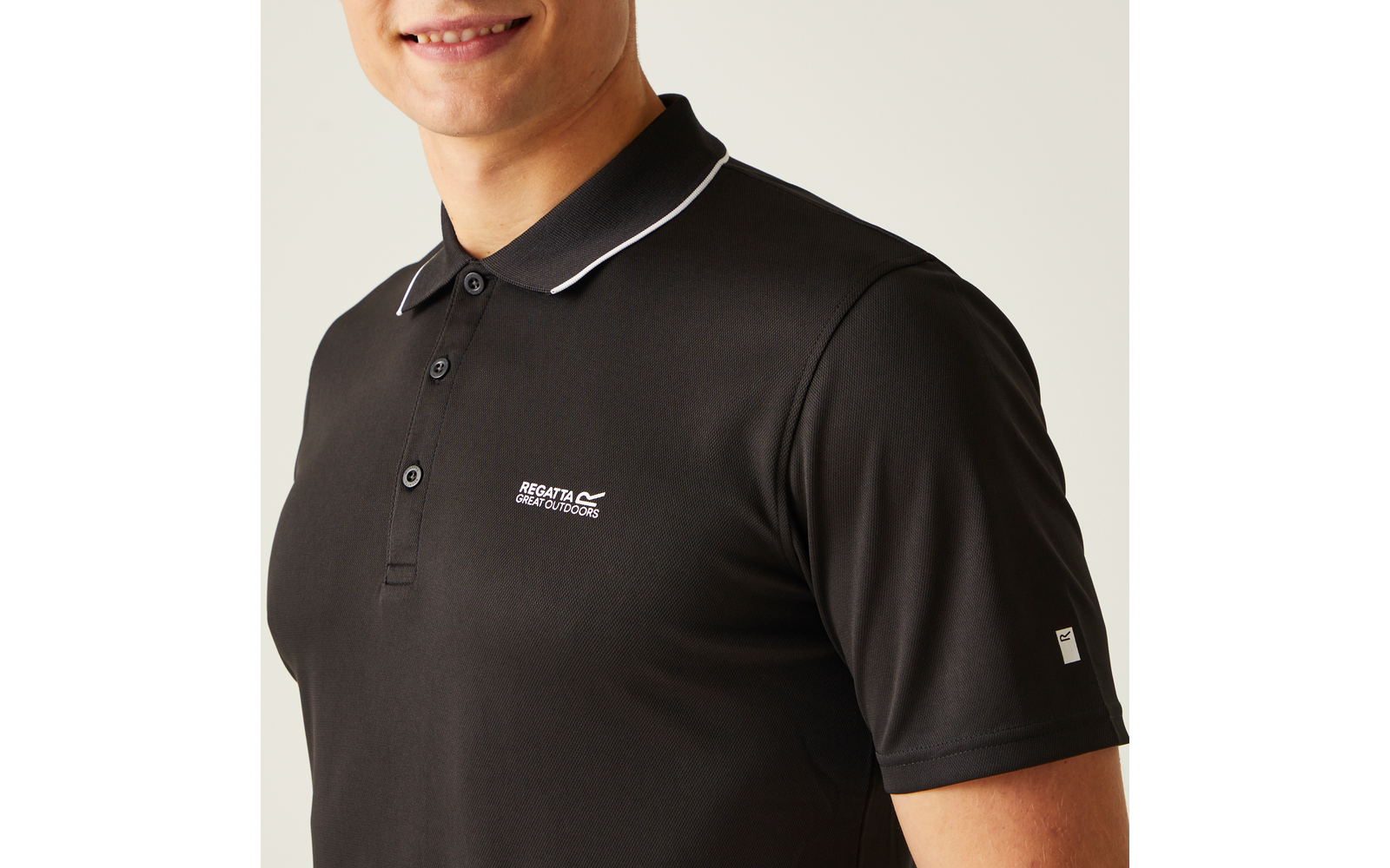 Oberkörper einer Person in schwarzem Polohemd mit weißer Kragenpaspel, drei Knöpfen, weißem Brustlogo und kleinem weißem Ärmellabel; teilweiser Gesichtsausschnitt mit Mund und Kinn