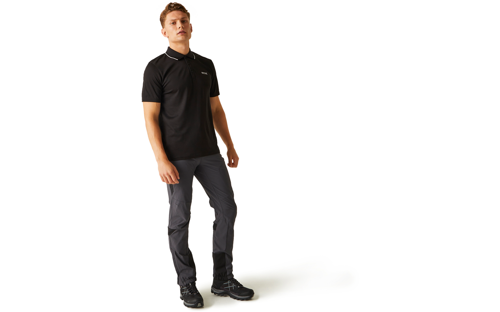 Ein Mann steht vor weißem Hintergrund, trägt ein schwarzes Poloshirt, graue Hosen und schwarze Schuhe